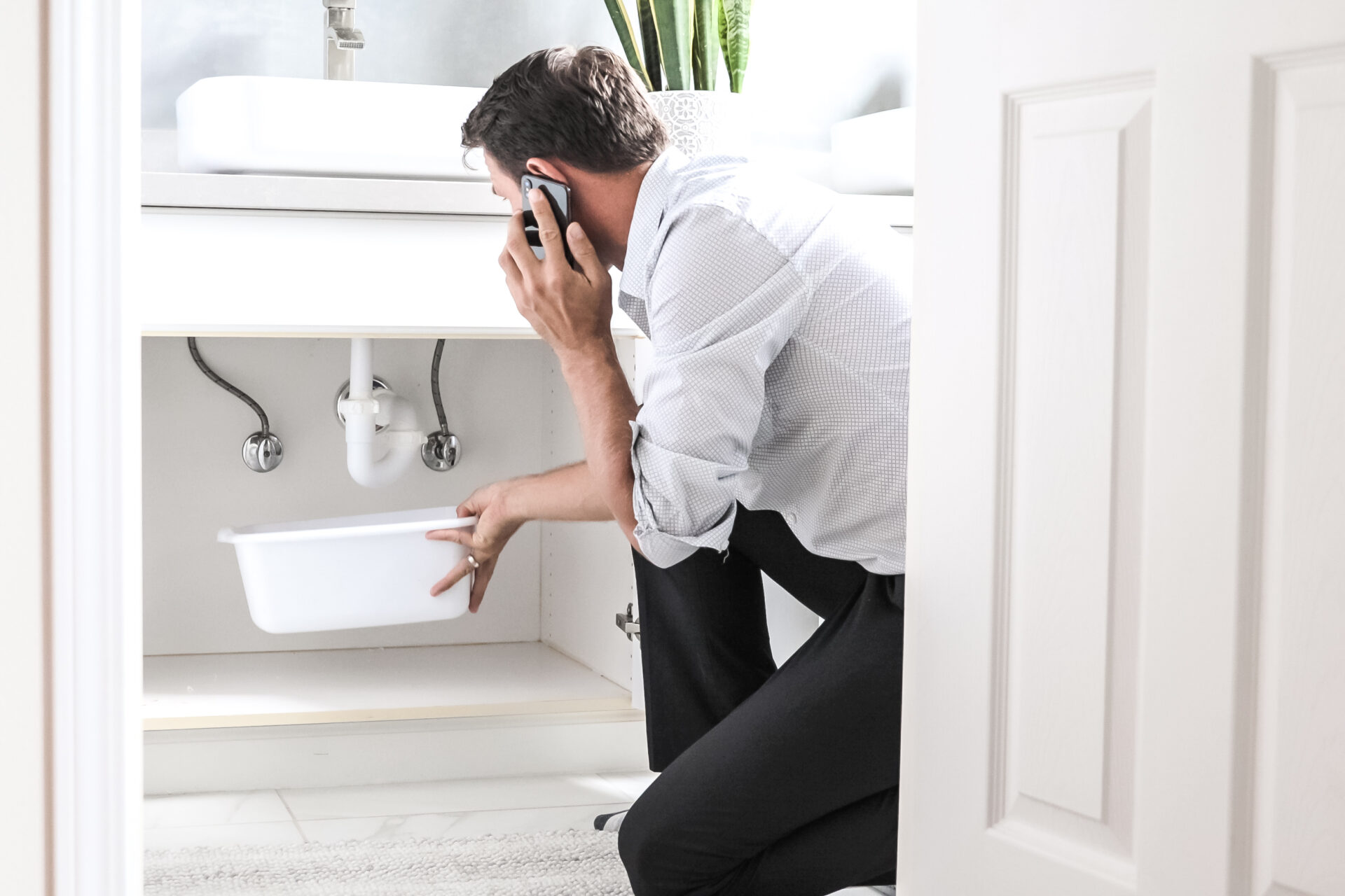 Sad Young Man Calling for Emergency Plumbing In Front Of Water Leaking From Sink Pipe